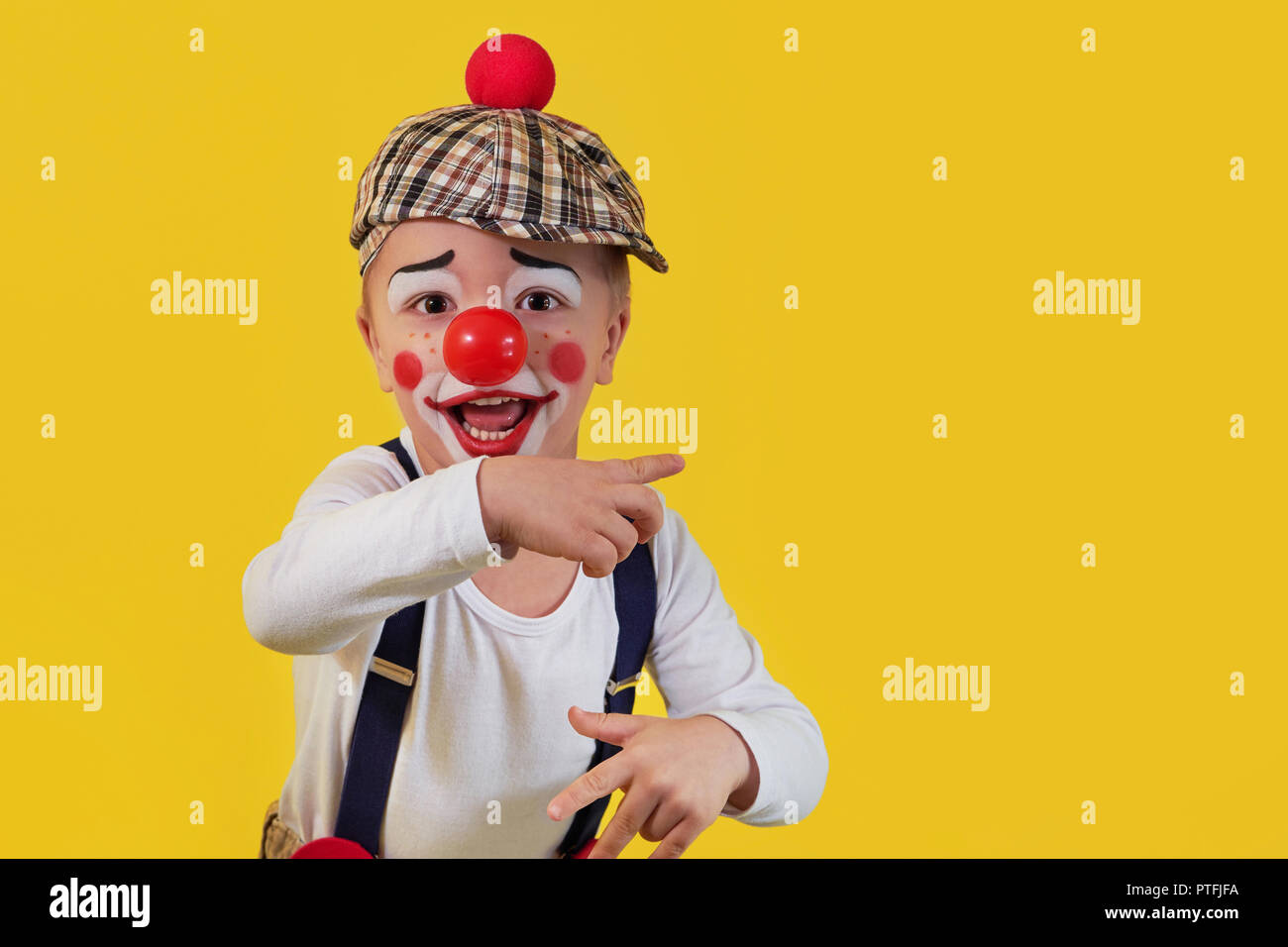Child clown smile shows a finger copy space area on yellow background. Funny little clown with red nose. Concept birthday, day 1 april, party. Beautiful portrait kid jester in studio. Stock Photo