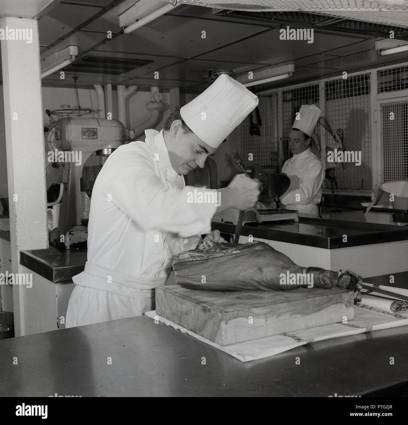 1950s, historical, male chef in uniform and tall hat or toque working in a kitchen below deck on a union-castle steamship, preparing a leg of serrano or cured ham on a wooden block. Stock Photo