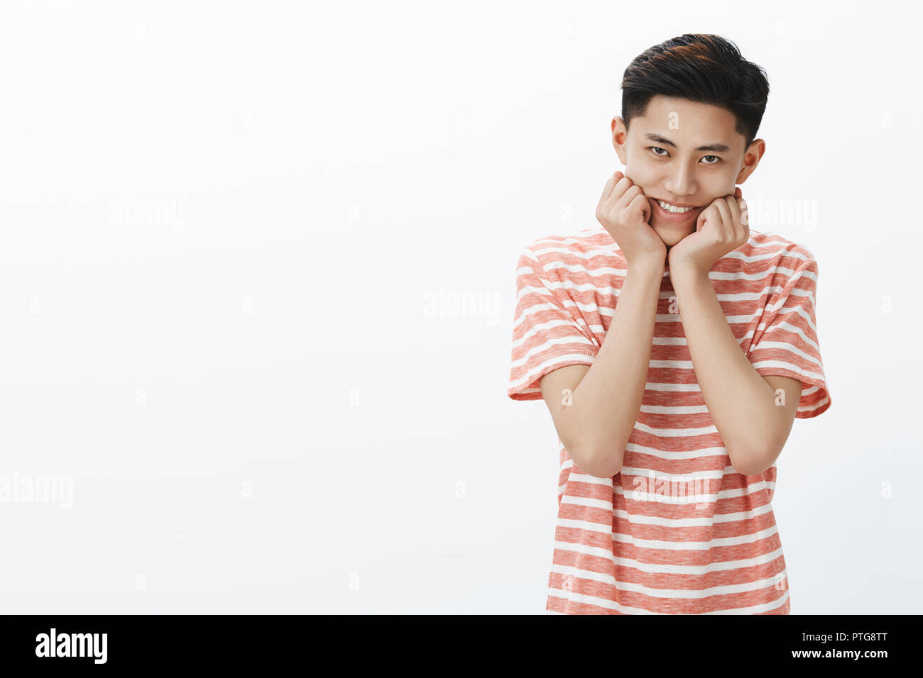 Portrait of silly and cute young adolescent male asian in striped t-shirt smiling leaning head on palms pressed to jawline trying look sweet and tender, gazing with admiration at camera over grey wall Stock Photo