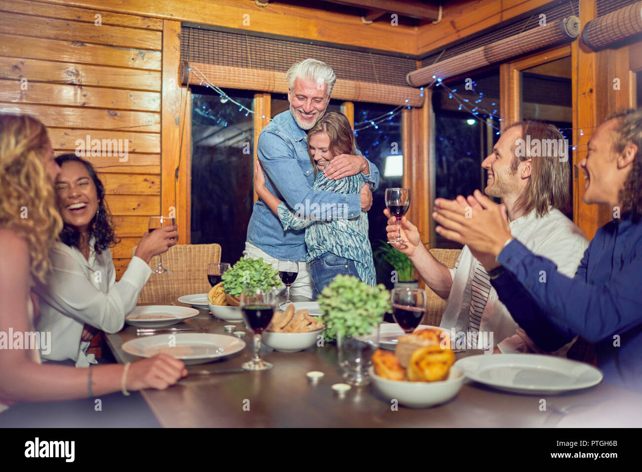 Friends clapping for affectionate couple hugging at dinner table Stock Photo
