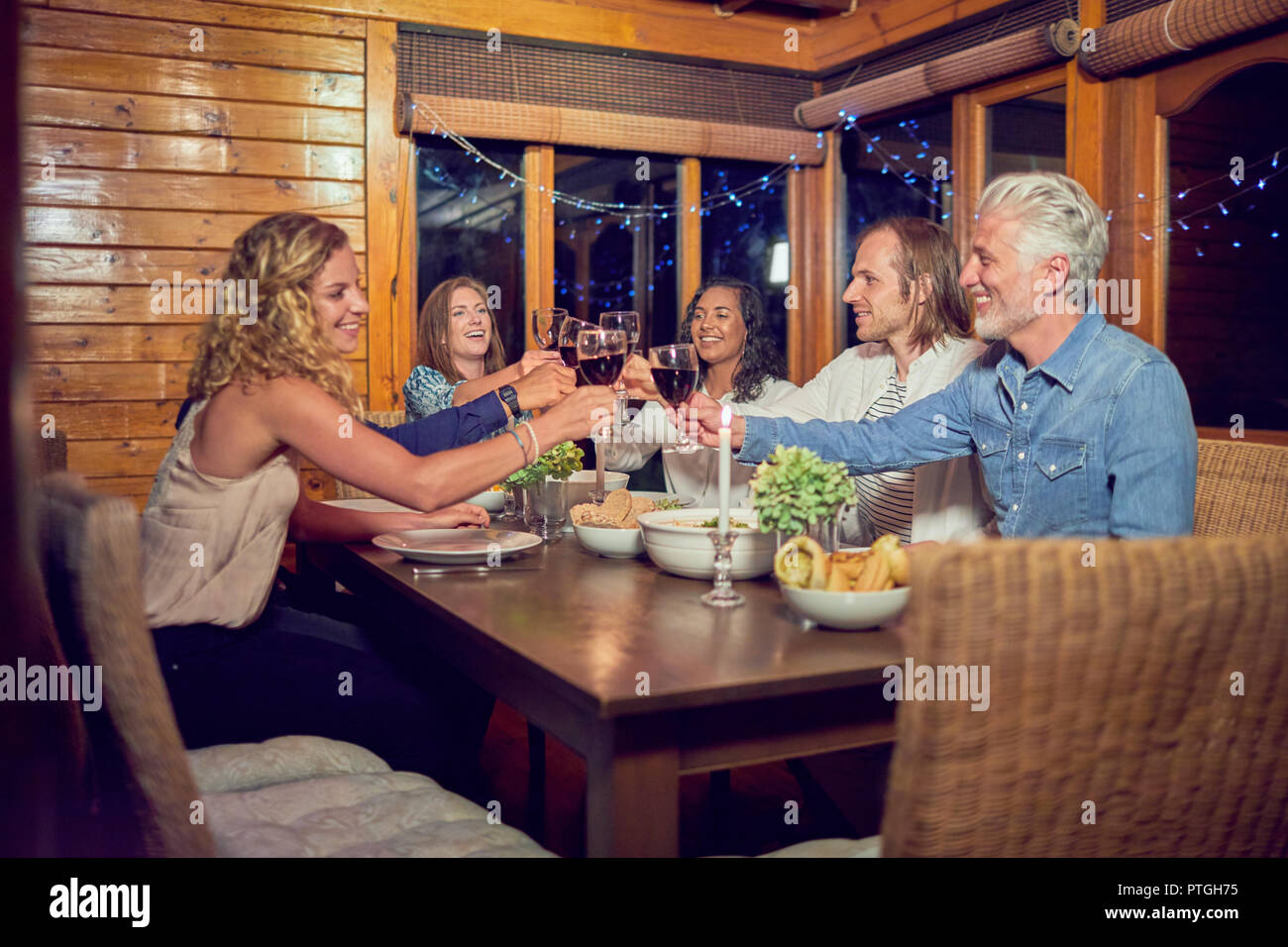 Friends toasting red wine glasses, enjoying dinner at cabin dining room table Stock Photo