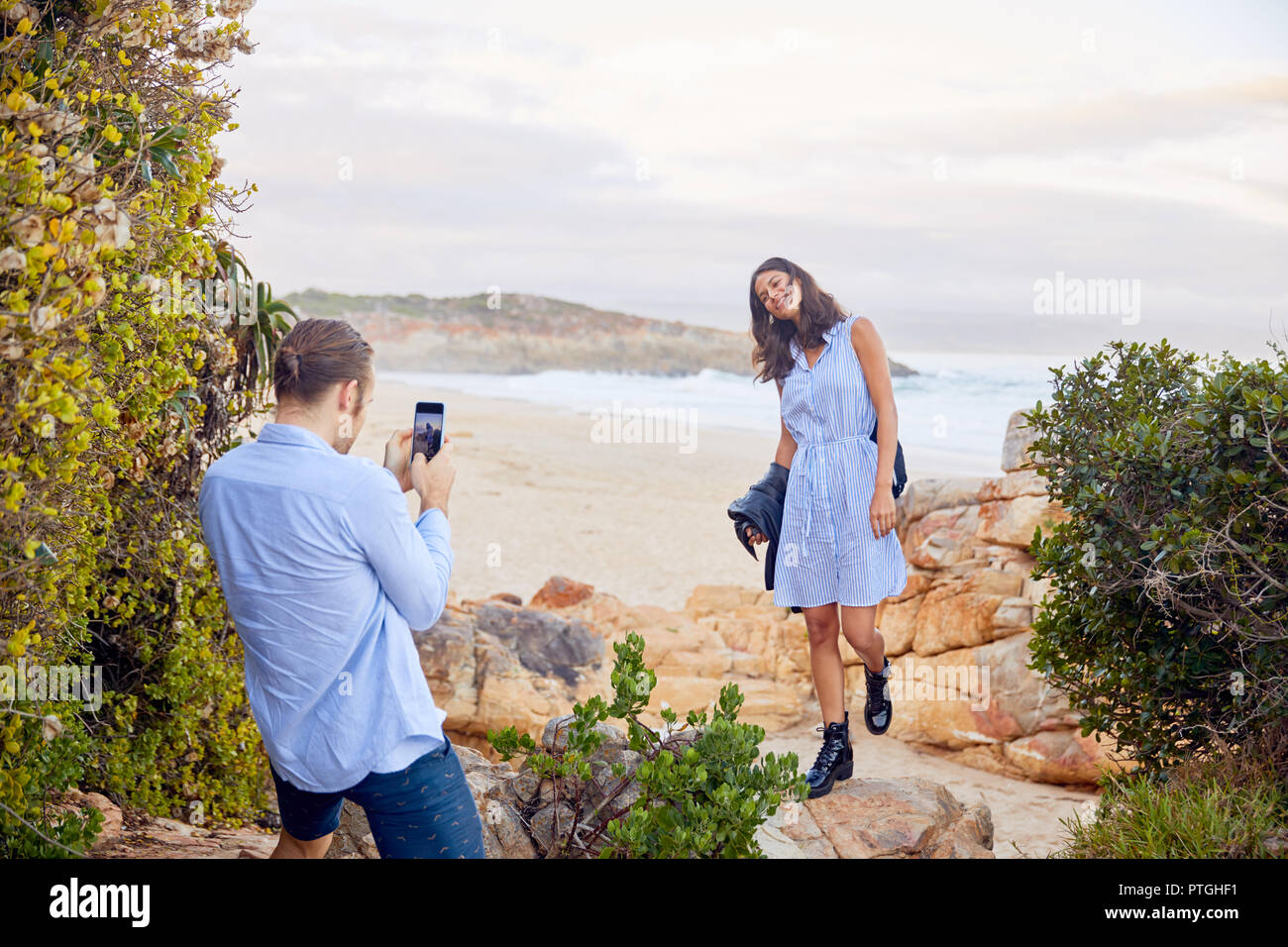 Young man with smart phone photographing girlfriend with ocean in background Stock Photo