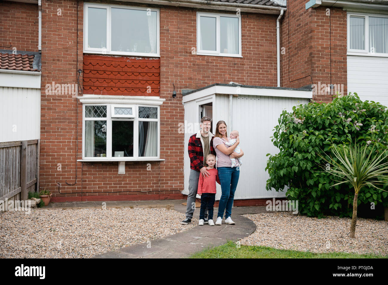 Family with two children standing and looking at the camera while smiling outside their house. They are proud owners of their home. Stock Photo