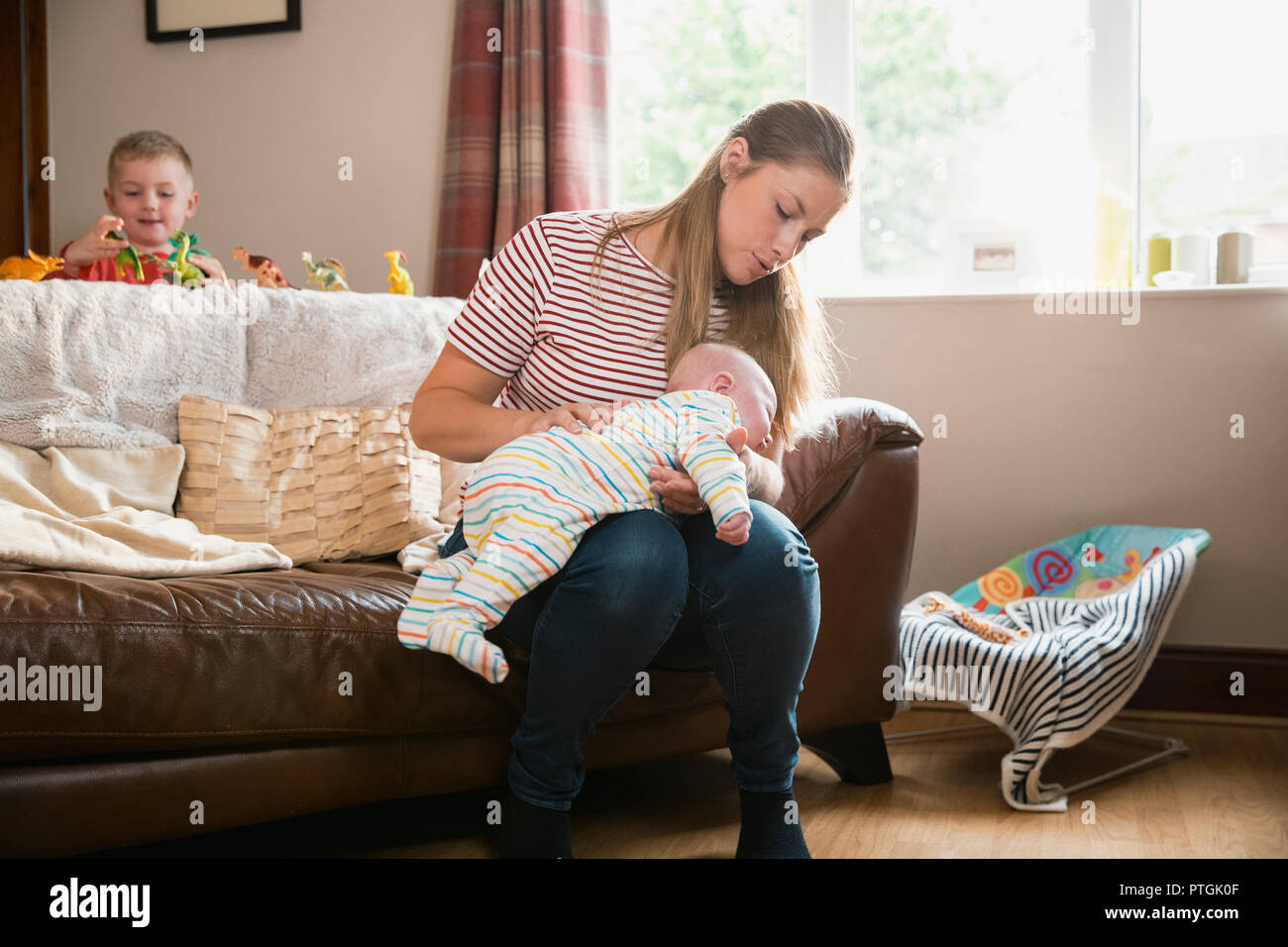 Mother sitting on the edge of the sofa in the living room and burping her newborn child after feeding him. Her other son is stadning behind the sofa p Stock Photo