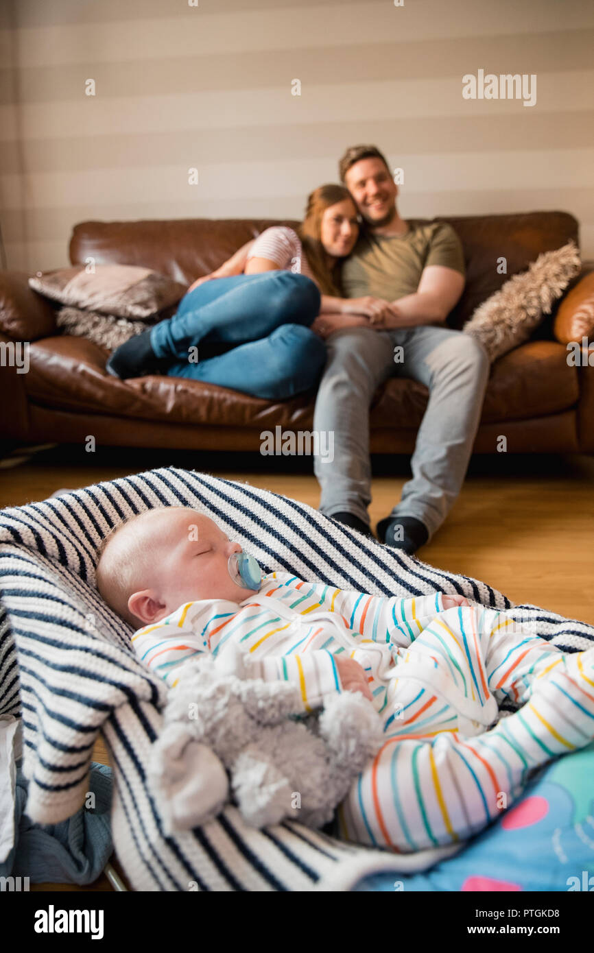Husband and wife sitting watching the tv and enjoying some peace and quiet while their newborn baby boy sleeps in a baby bouncer. Stock Photo