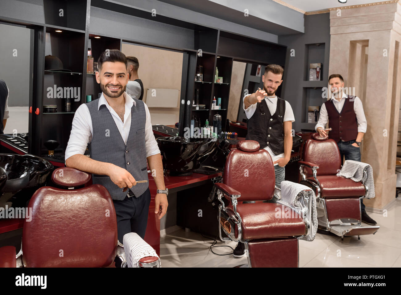 Three professional barbers standing near hairdresser chairs and posing. Handsome, confident masters looking at camera and holding scissors in hands. Interior of modern barbershop. Stock Photo
