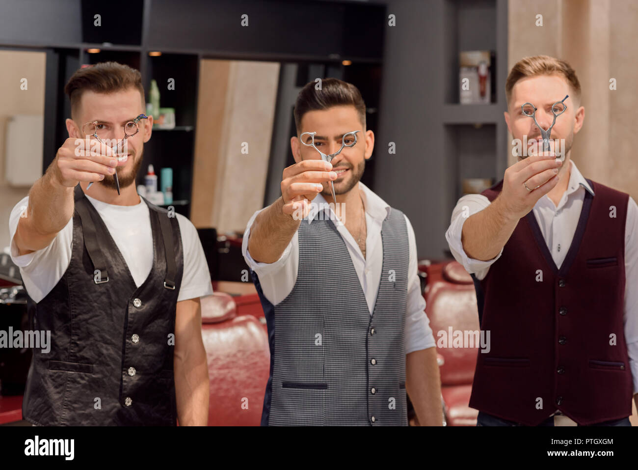 Positive barbers showing and holding scissors, and looking at camera through them. Professional masters standing and posing in barbershop. Handsome hairdressers wearing white shirts and vests. Stock Photo