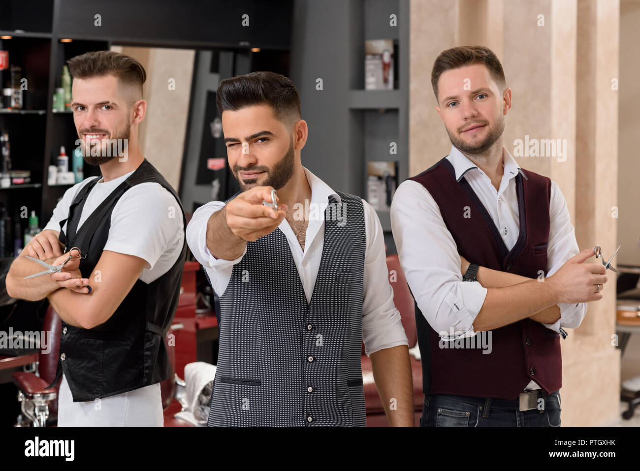 Three handsome hairstylists posing and standing in barbershop. Charming bearded master pointing with scissors at camera and winking. Masters looking at camera and holding instruments for hairstyling. Stock Photo