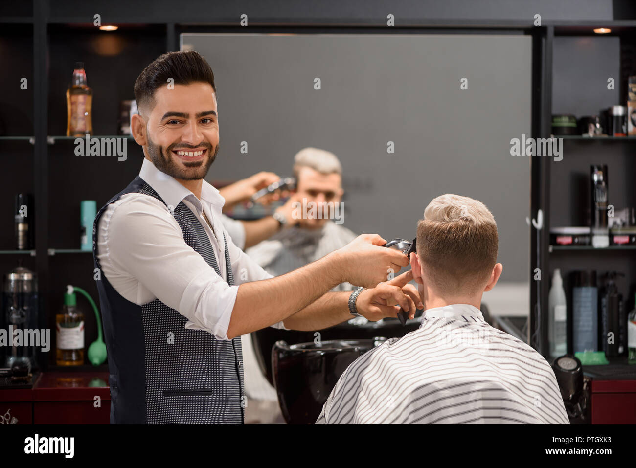 Smiling hairstylist working with client in barbershop. Bearded barber looking at camera and trimming stylish haircut of young man. Reflection in mirror of client covered with coiffure cape. Stock Photo