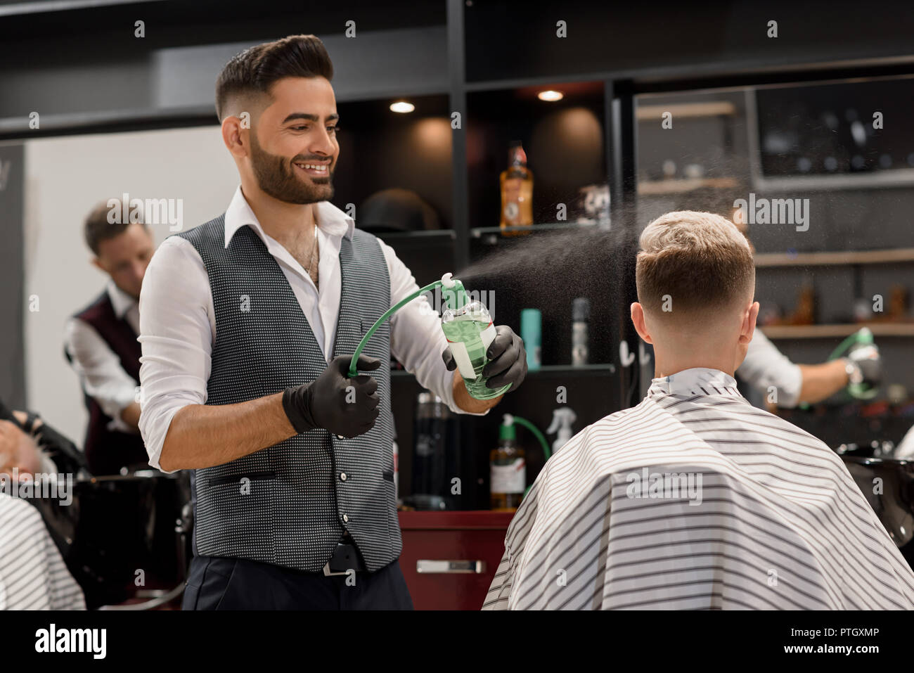 Smiling hairdresser sprinkling water on client's hair. Barber using sprayer bottle with green liquid. Master wearing classic white shirt and grey vest, while client sitting in striped haircut gown. Stock Photo