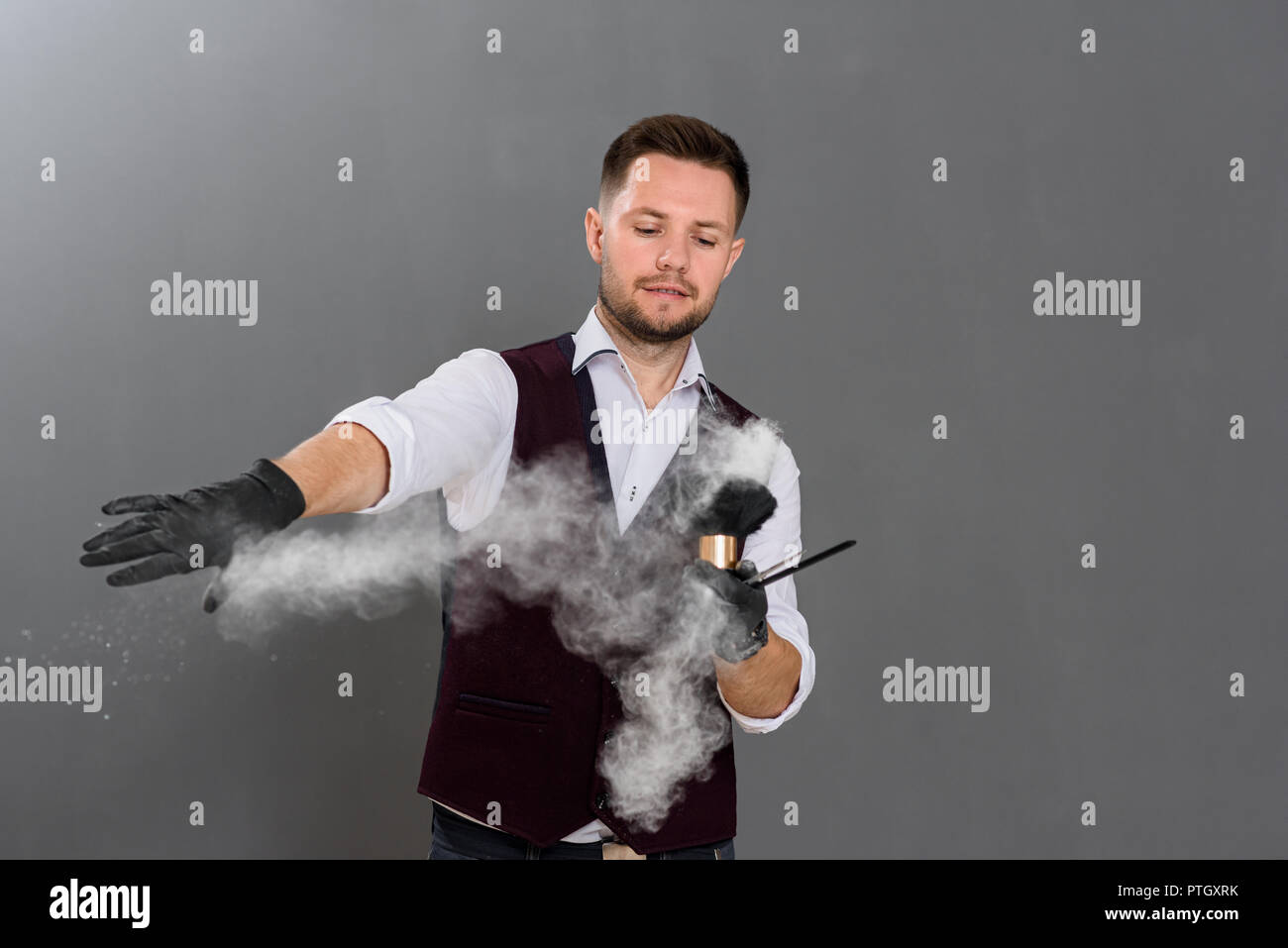 Handsome barber posing in studio on grey background. Male hairdresser  holding instruments for haircut and brush, he spilling powder in air.  Master wearing white classic shirt and vest Stock Photo - Alamy