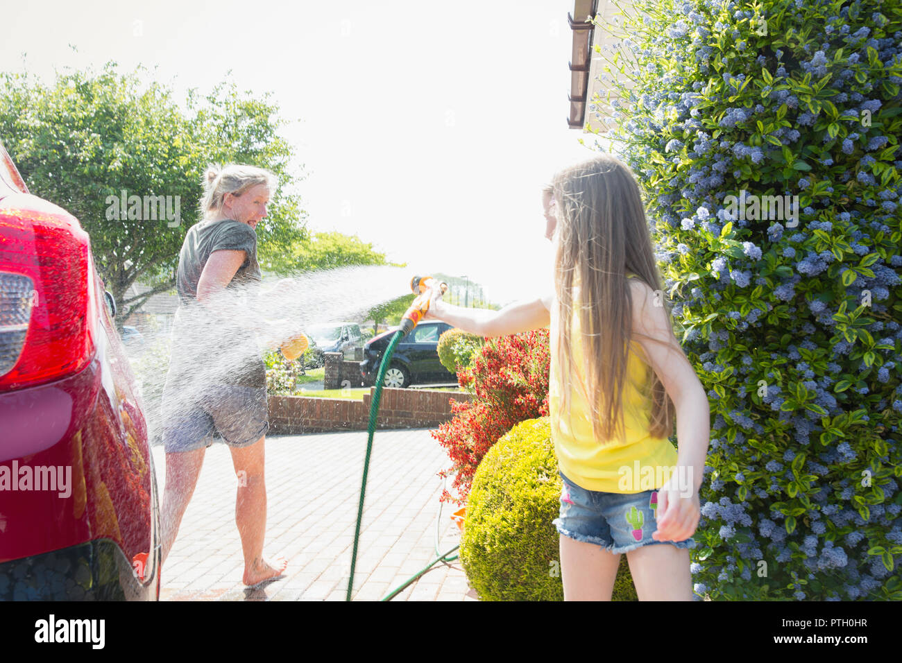 Playful daughter spraying mother with hose in sunny driveway Stock Photo
