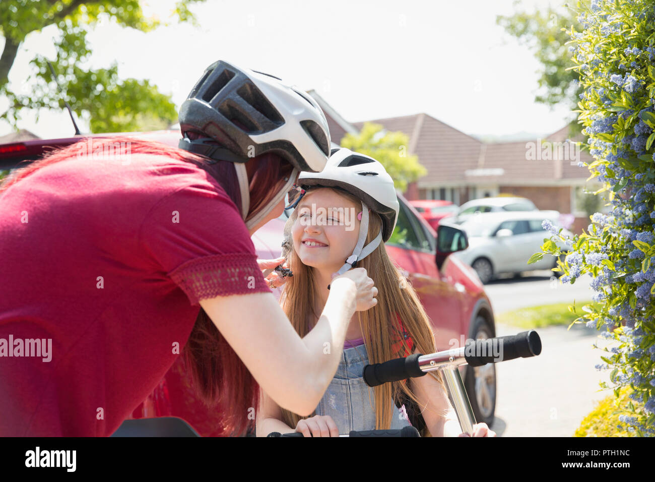 Mother fastening helmet on daughter riding scooter in sunny driveway Stock Photo