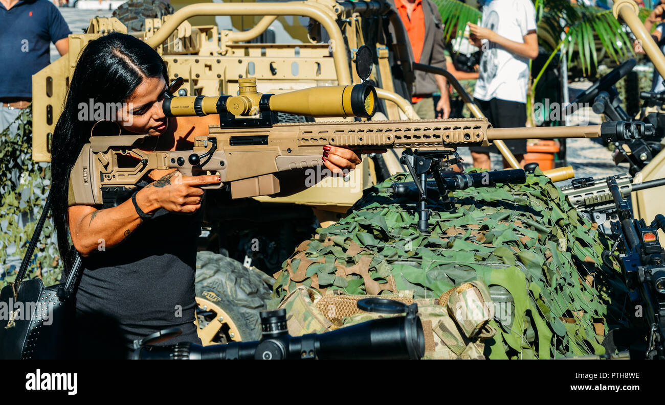 Young beautiful feminine civilian with handbag and tattoos aiming using a high-tech army sniper rifle Stock Photo