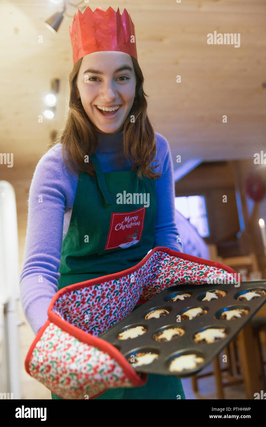 Portrait enthusiastic teenage girl in Christmas apron and paper crown baking muffins Stock Photo