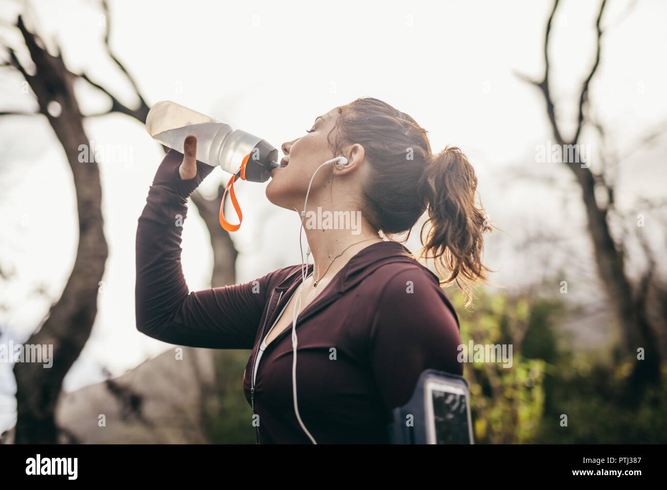Woman drinking water after a running exercise outdoors. Female runner taking a break and drinking water from bottle in morning. Stock Photo