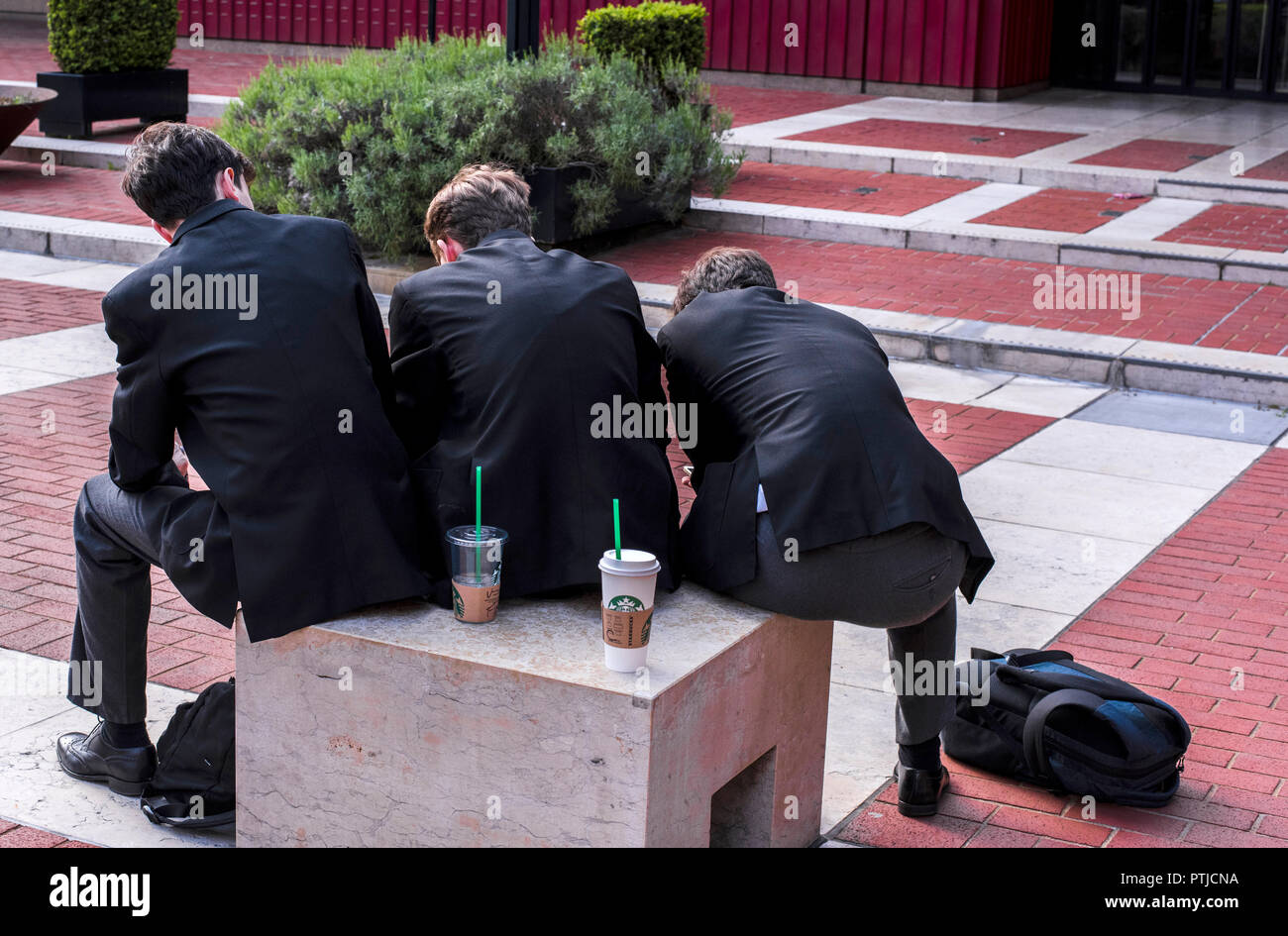 3 boys in their school uniform and a rucksack on the floor are seen from behind creating a strange sloping shape in the gardens of the British Library. Stock Photo