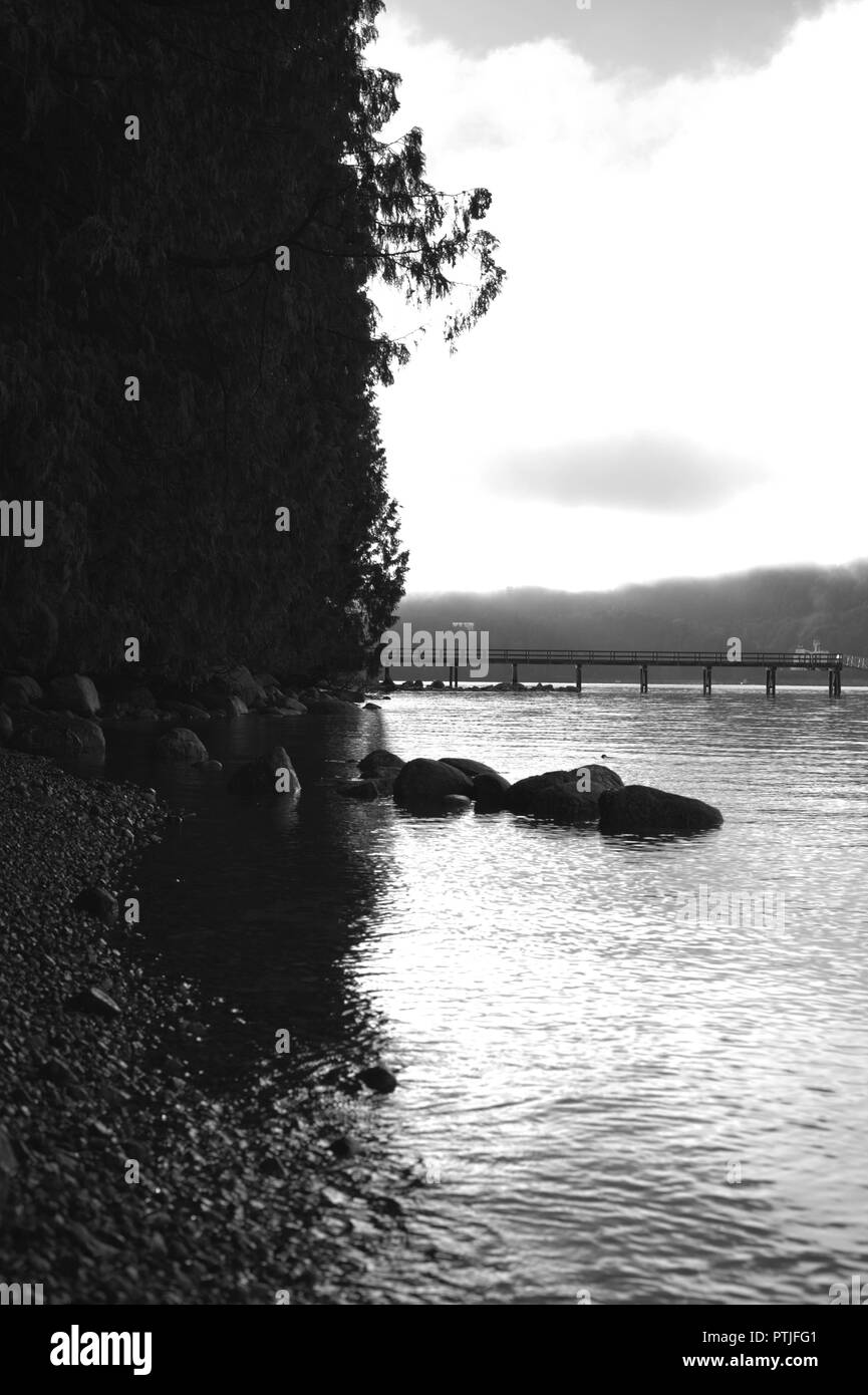 Black and white landscape of the Indian arm river, with the rocky shore and Vancouver city mountains in the background, with a bridge in the horizon Stock Photo