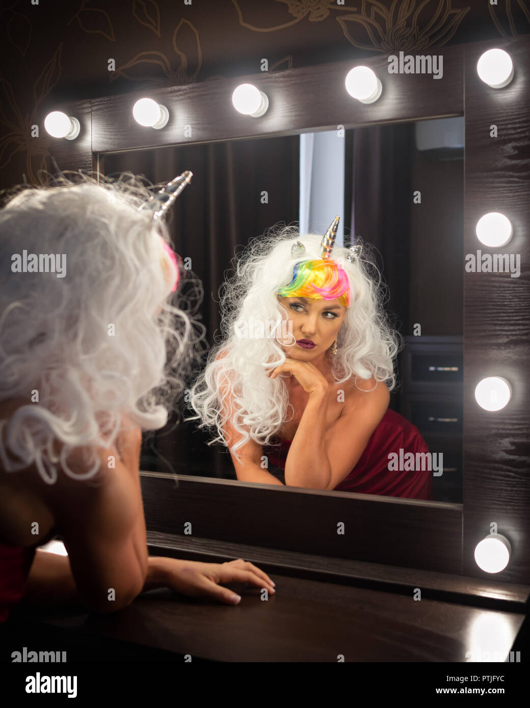 Freaky young woman in unusual wig looks at himself in the mirror in dressing room. Portrait of strange lady in red dress. Serious girl unicorn. Stock Photo