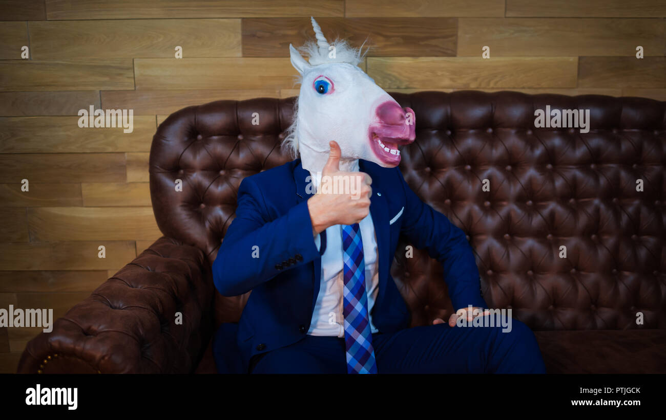 Funny unicorn in elegant suit sits on sofa like a boss and showing gesture thumbs up. Portrait of unusual man at home. Freaky young manager in mask Stock Photo