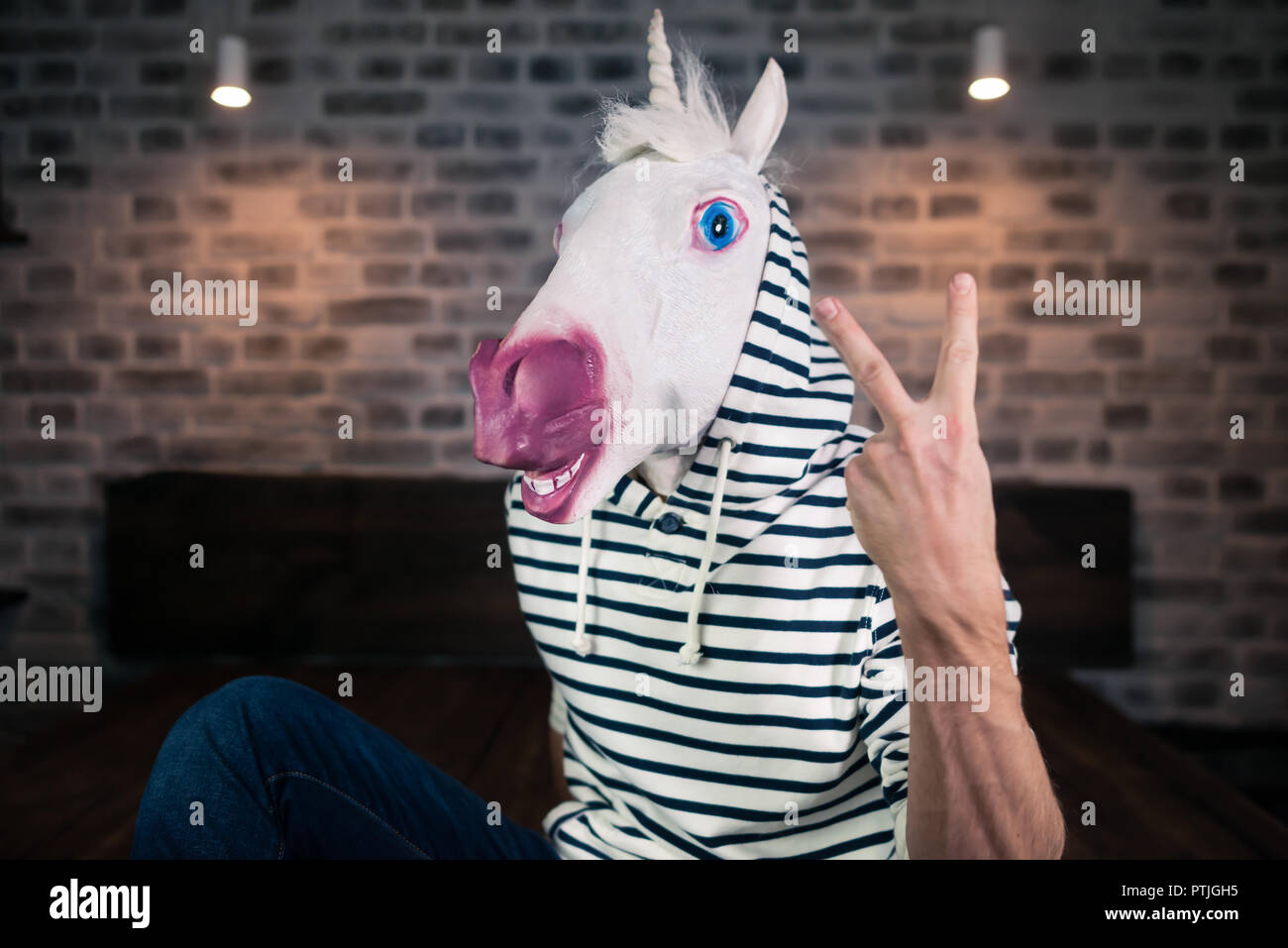Freaky young man in comical mask sits on bed in stylish apartment. Funny unicorn in casual clothes is showing two fingers up gesture. Stock Photo