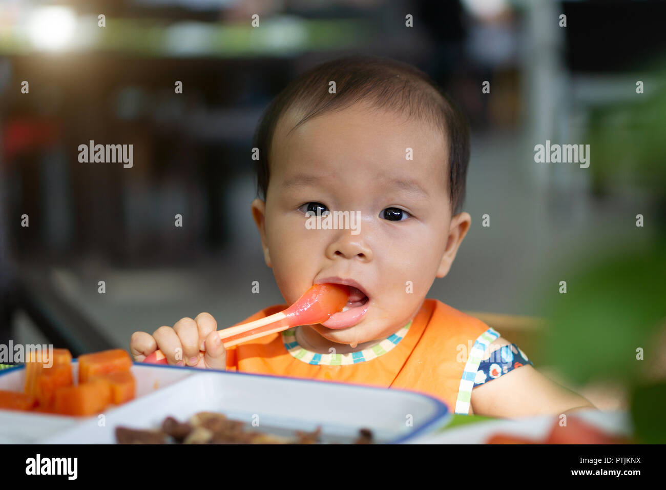 Asian baby girl in restaurant with a spoon eats food. Stock Photo