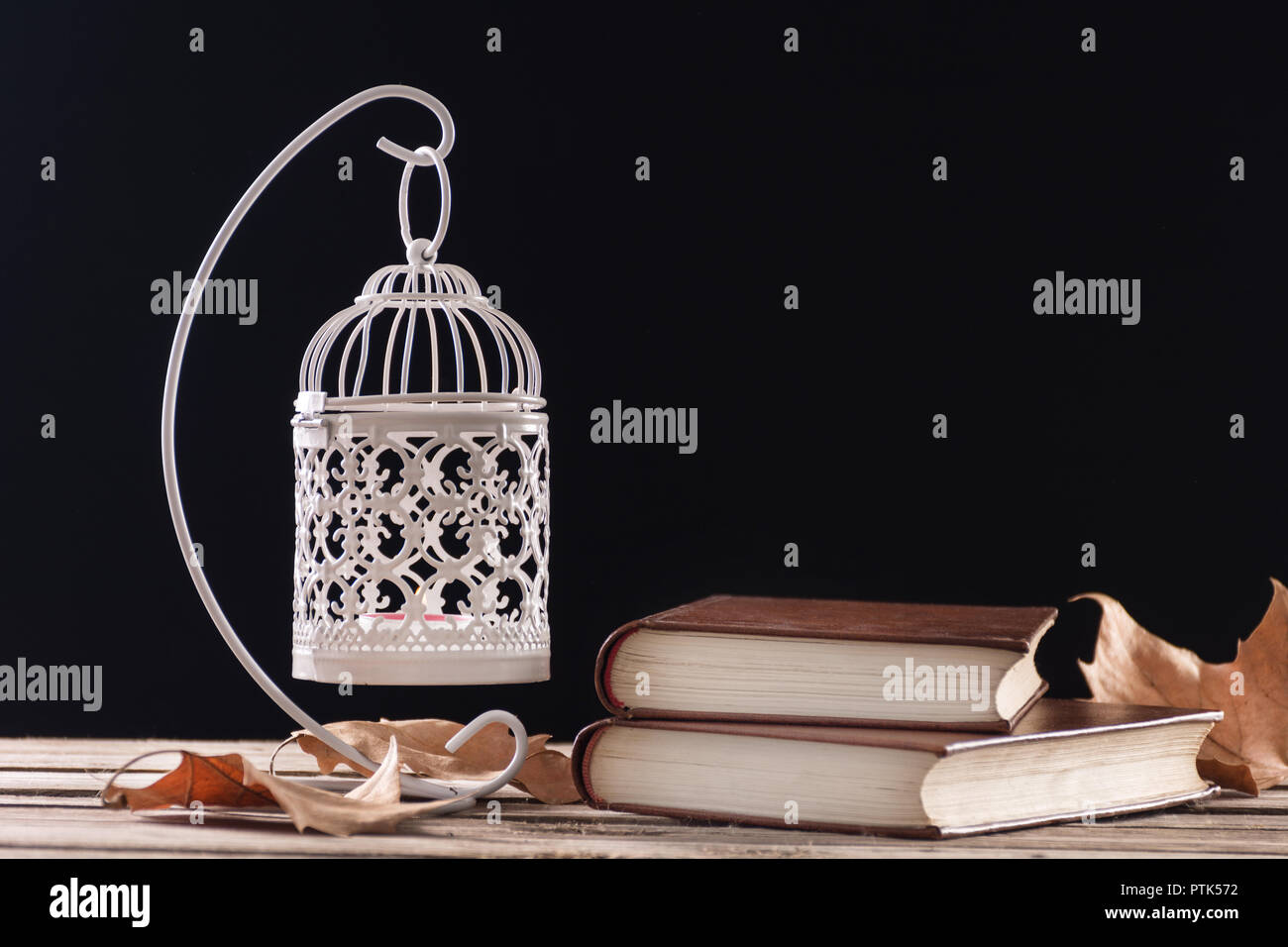 Retro decorative cage with candle and old books on wooden desk with fallen dry leaf, black background. Education and autumn concept. Close up Stock Photo