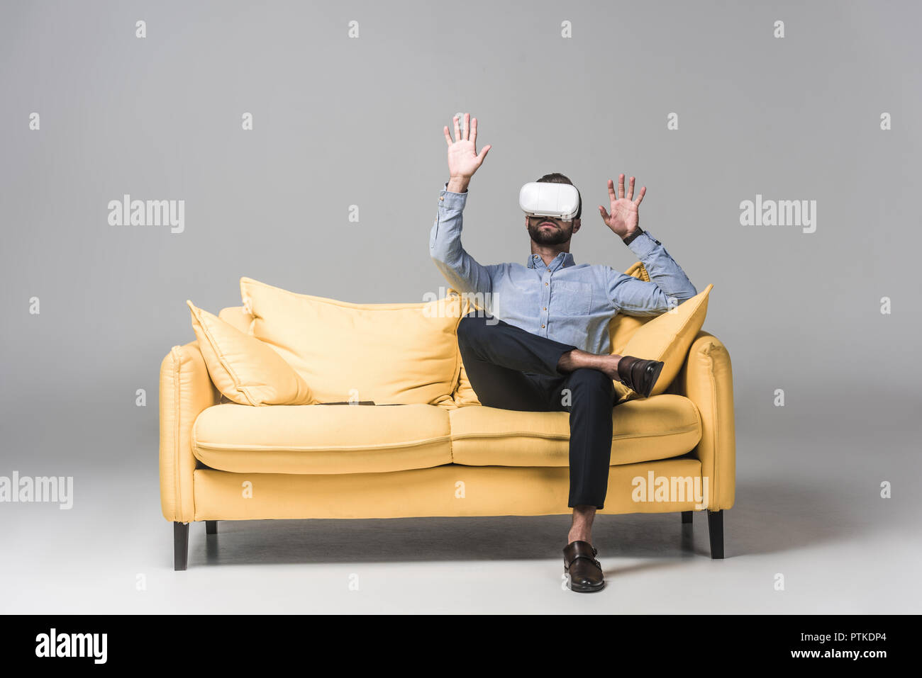 bearded man gesturing and using virtual reality headset and sitting on yellow sofa on grey Stock Photo