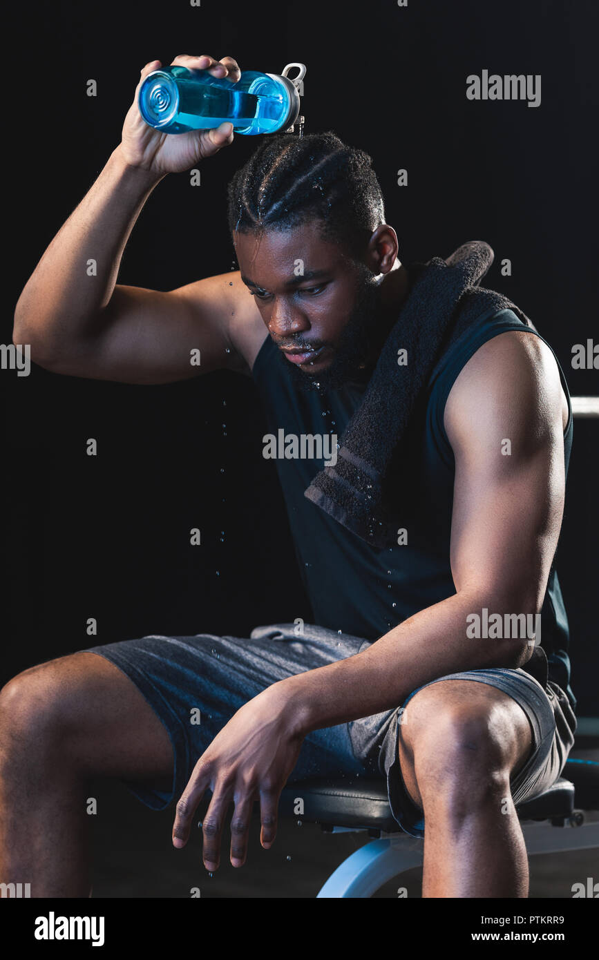 tired african american sportsman pouring water on head while sitting after workout in gym Stock Photo