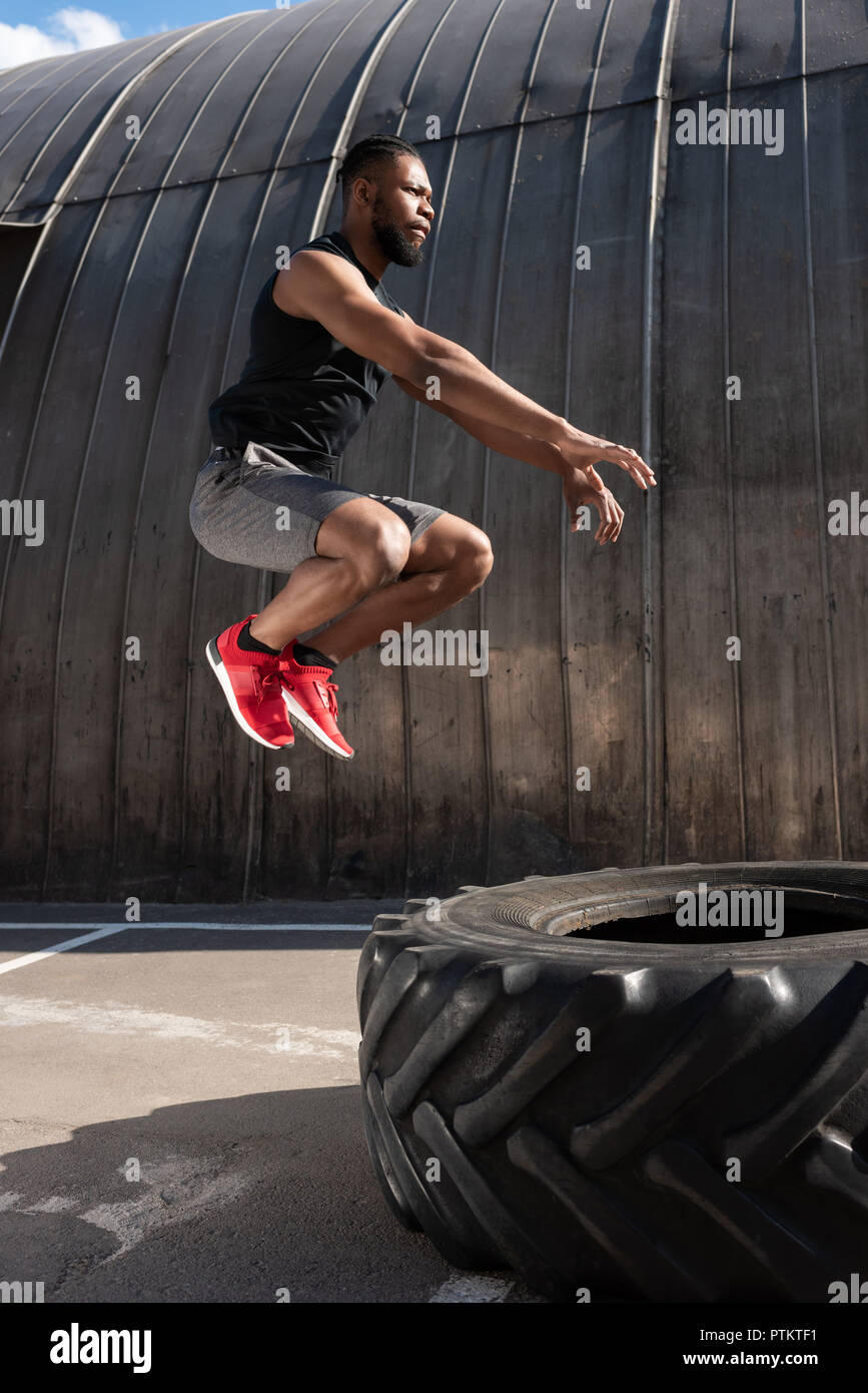 side view of muscular african american man jumping while training with tyre on street Stock Photo