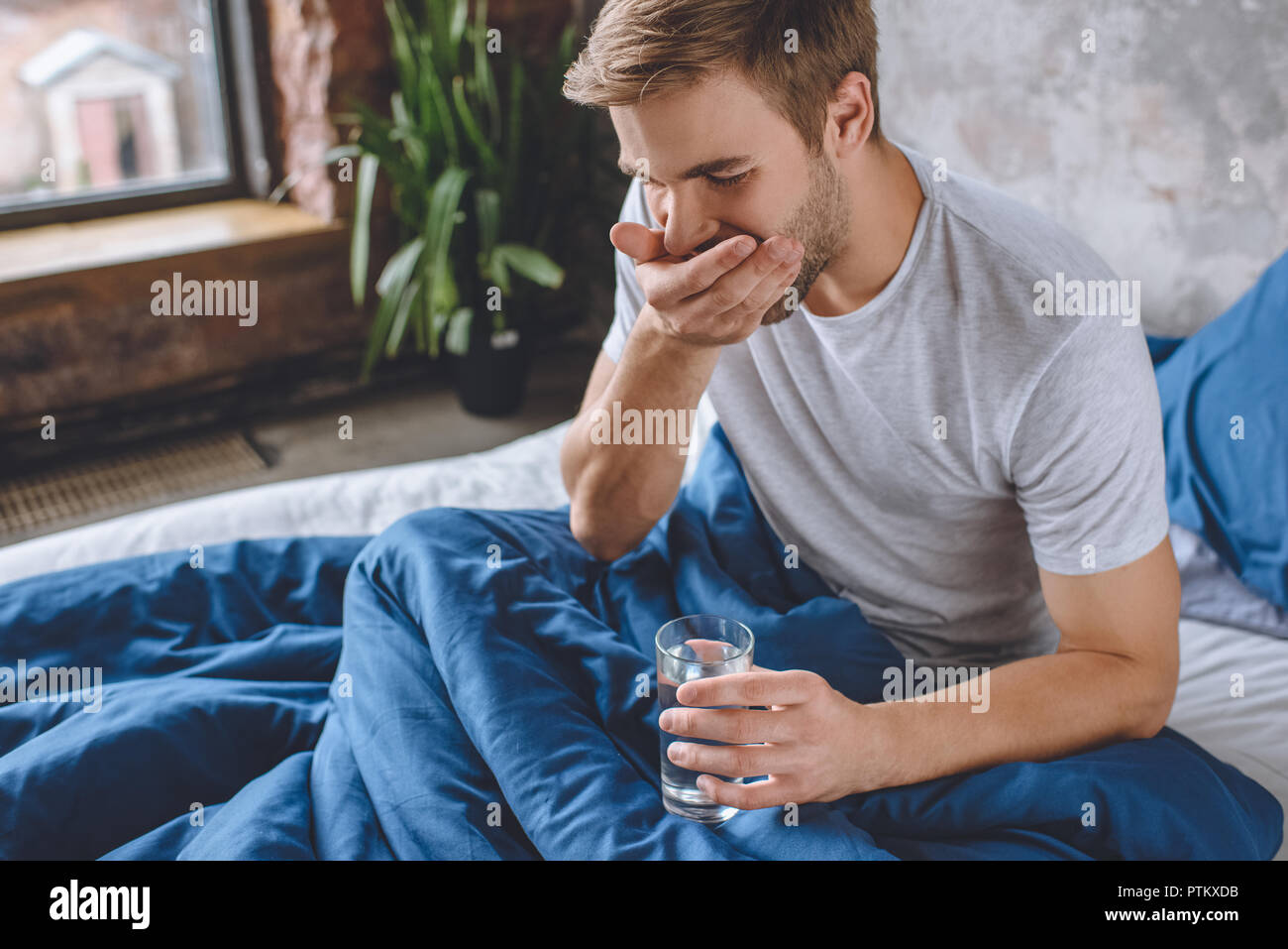 selective focus of young man taking pill and holding glass of water in bed at home Stock Photo