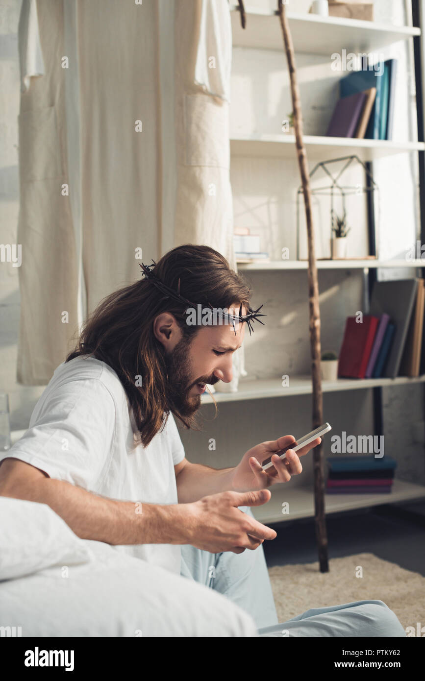 side view of Jesus in crown of thorns using smartphone in bedroom at home Stock Photo