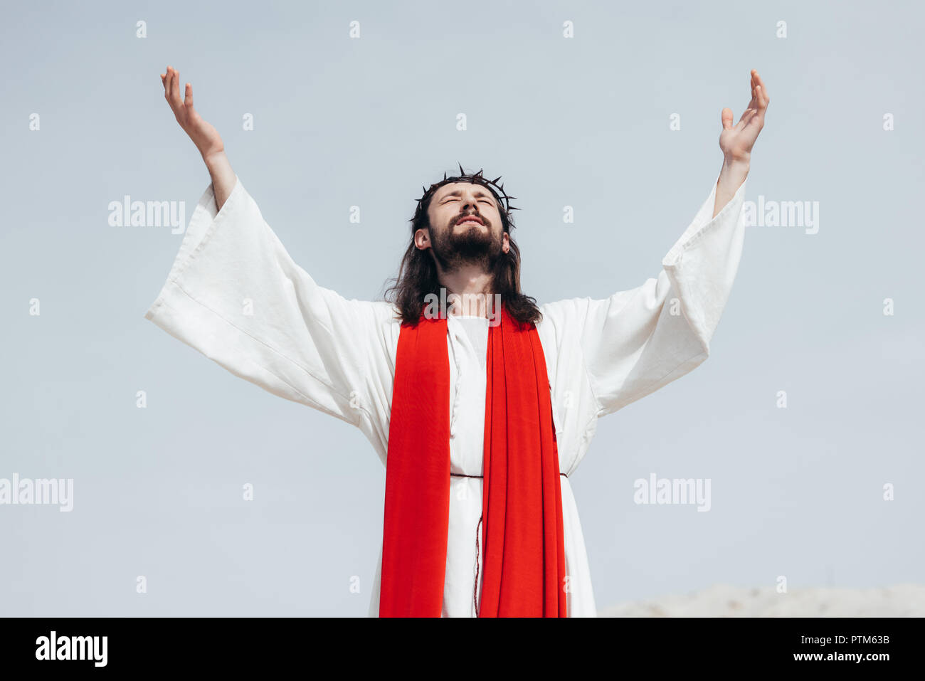 Jesus in robe, red sash and crown of thorns standing with raised hands and praying in desert Stock Photo