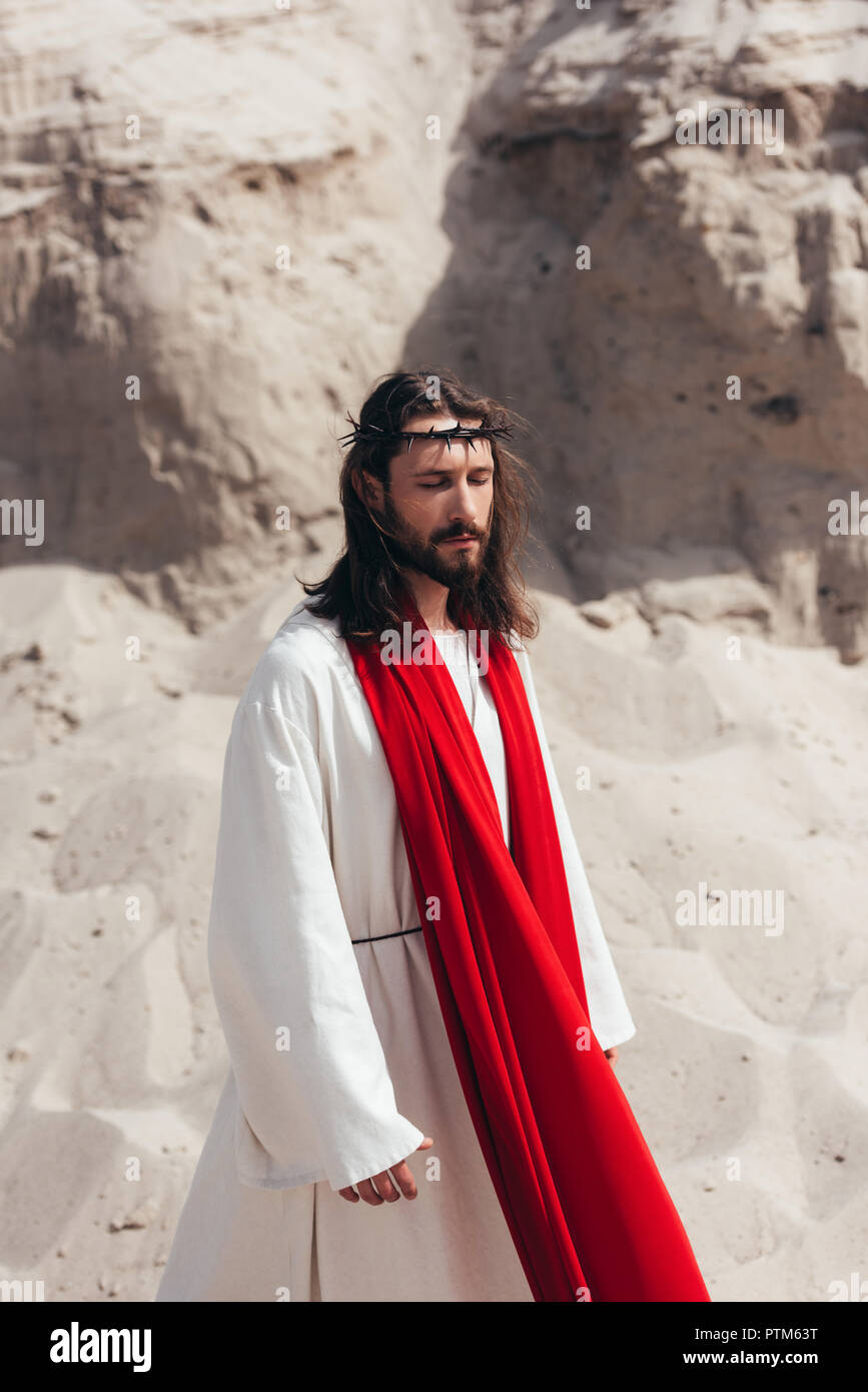 handsome Jesus in robe, red sash and crown of thorns walking with closed eyes in desert Stock Photo