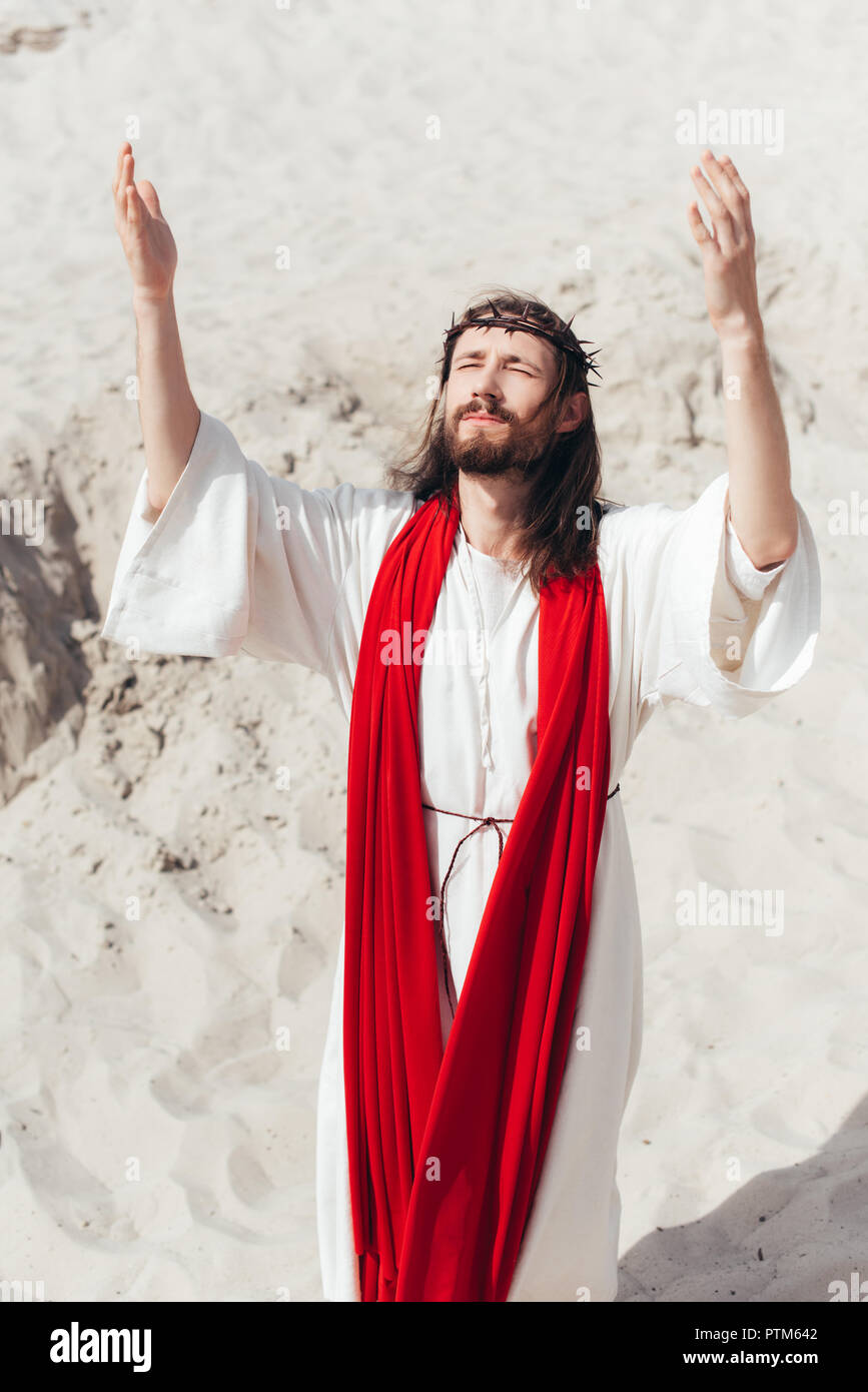 Jesus in robe, red sash and crown of thorns standing with raised hands and praying in desert Stock Photo