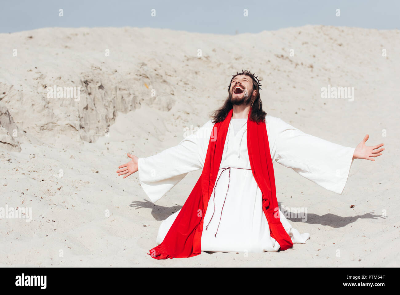 laughing Jesus in robe, red sash and crown of thorns standing on knees with open arms in desert Stock Photo