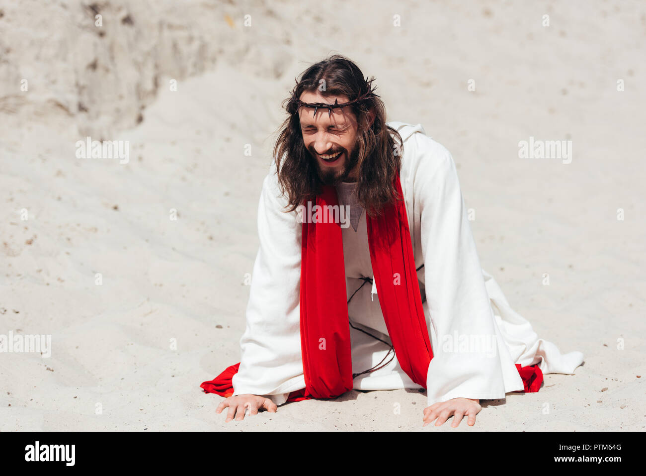 laughing Jesus in robe, red sash and crown of thorns standing on knees and touching sand with hands in desert Stock Photo
