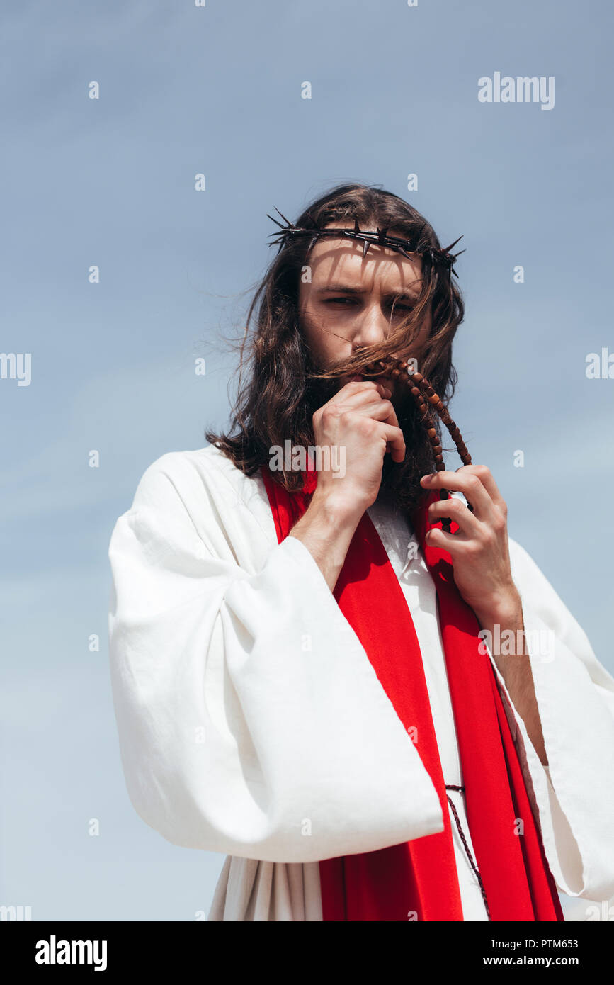 Jesus in robe, red sash and crown of thorns kissing rosary against blue sky Stock Photo