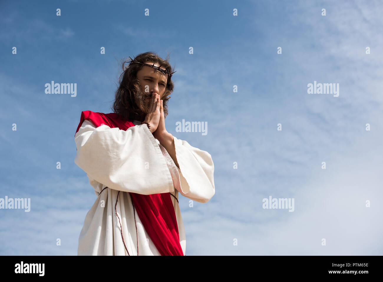 low angle view of Jesus in robe and red sash praying against blue sky Stock Photo
