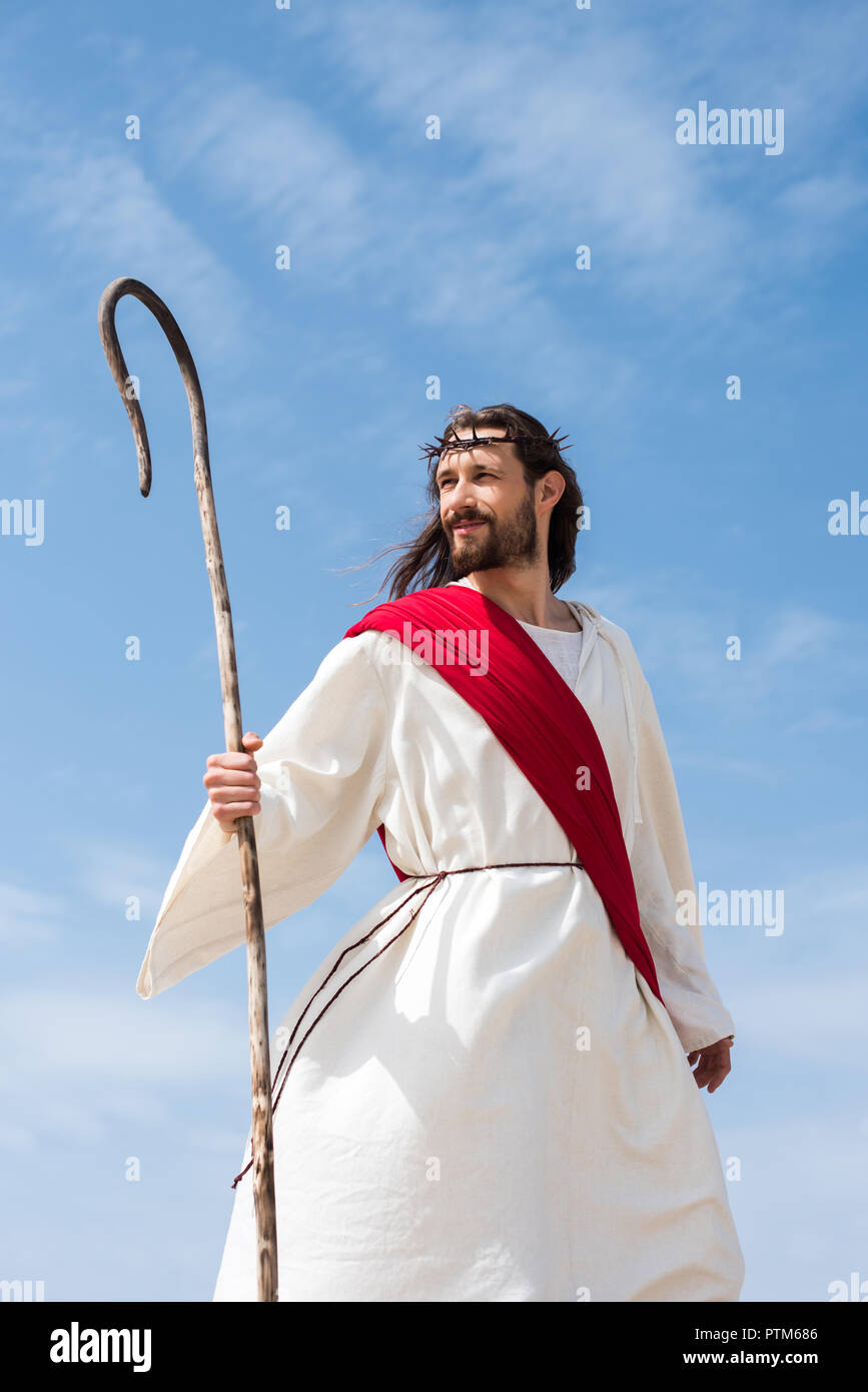 cheerful Jesus in robe, red sash and crown of thorns standing with wooden staff in desert Stock Photo