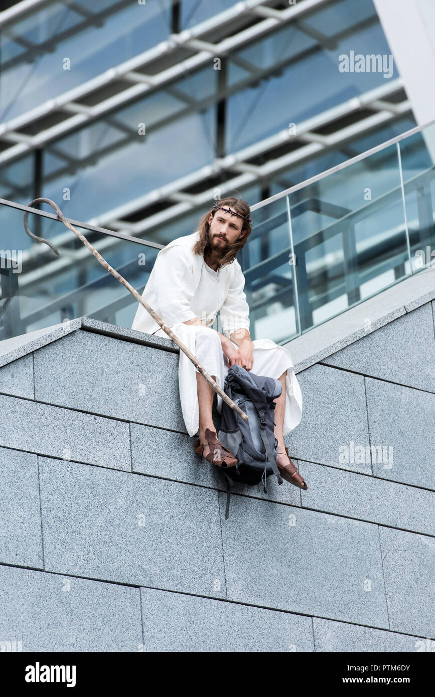 low angle view of Jesus in robe and crown of thorns sitting with travel bag and staff on staircase side Stock Photo