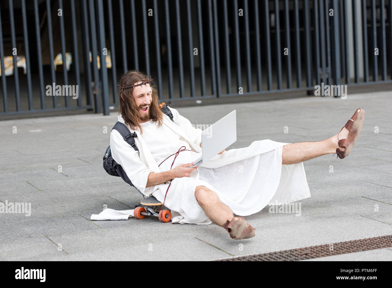 laughing Jesus in robe and crown of thorns sitting on skateboard and using laptop in city Stock Photo