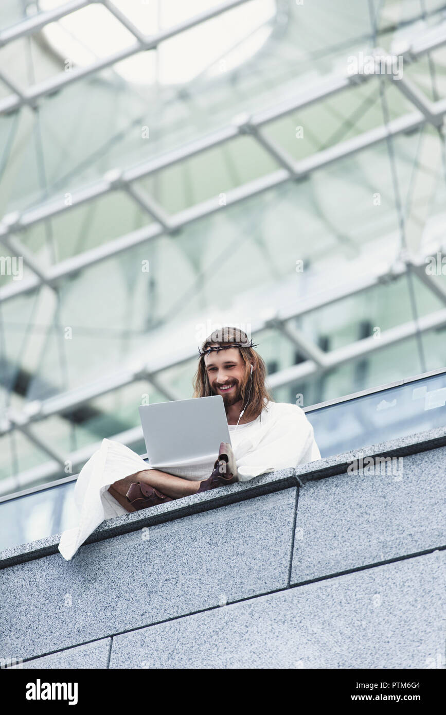 low angle view of laughing Jesus in robe and crown of thorns using laptop while sitting on wall Stock Photo