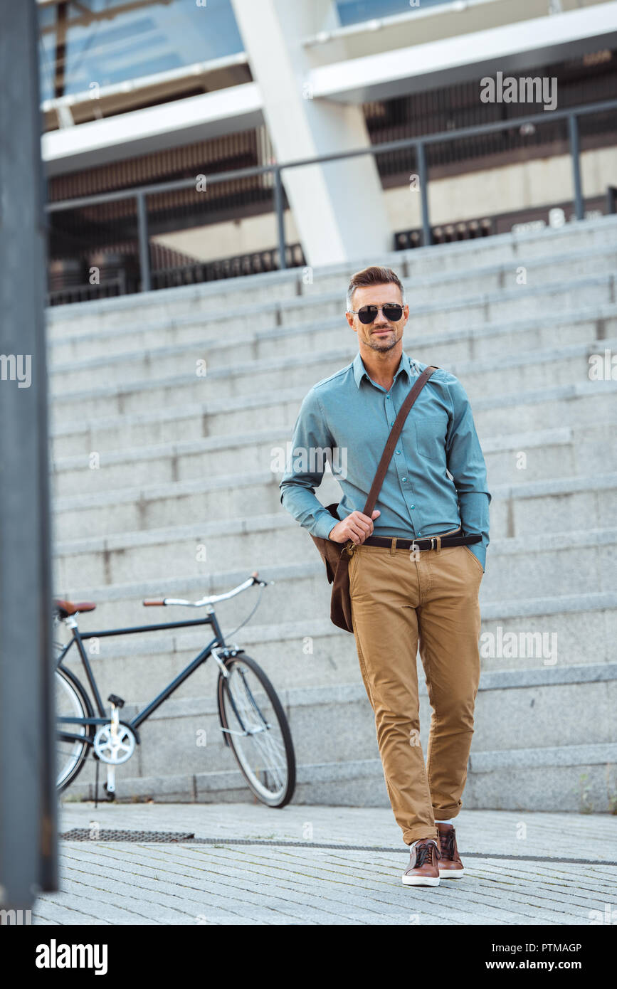handsome middle aged man in sunglasses smiling at camera while walking on street, bicycle behind Stock Photo