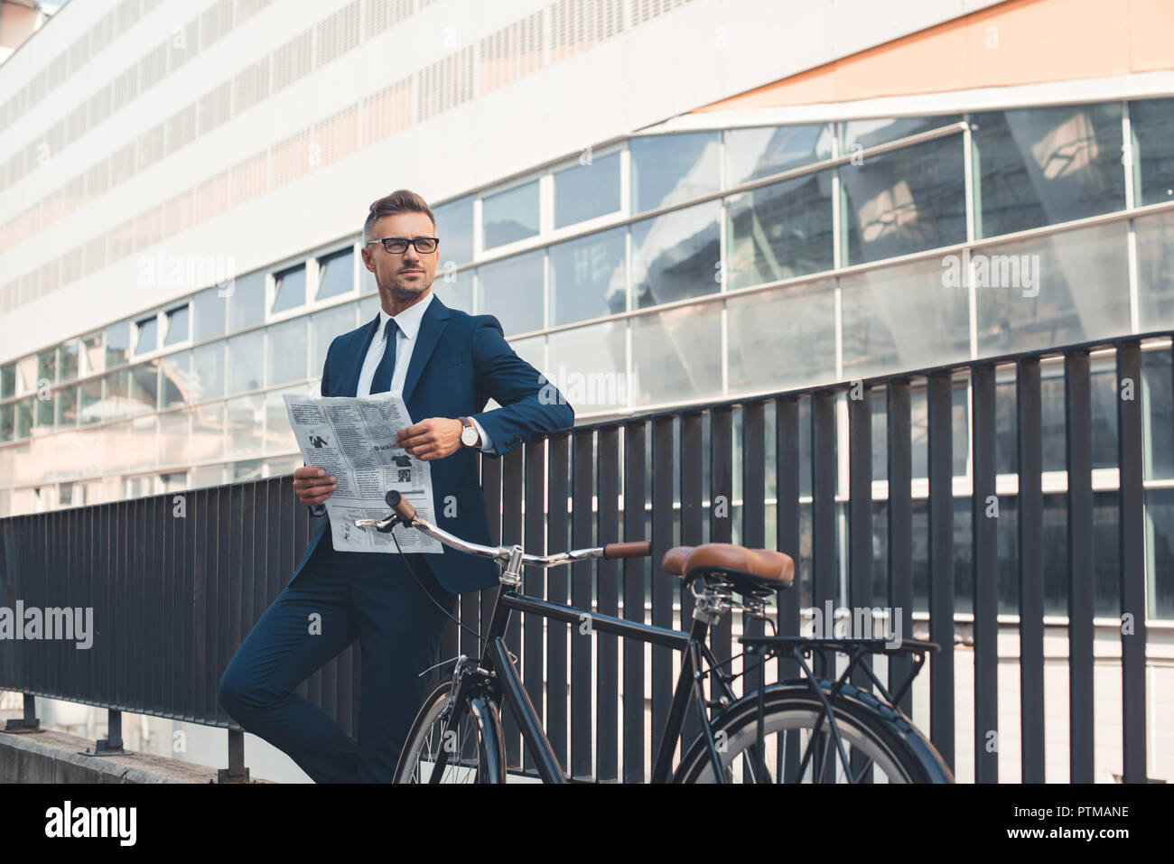 successful businessman in formal wear holding newspaper and looking away while standing with bicycle on street Stock Photo