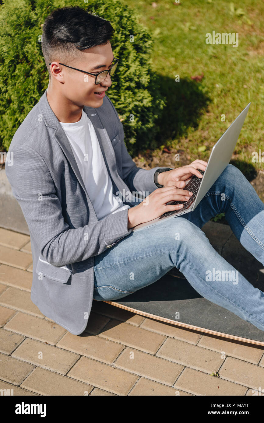 asian teleworker using laptop while sitting on longboard on street Stock Photo