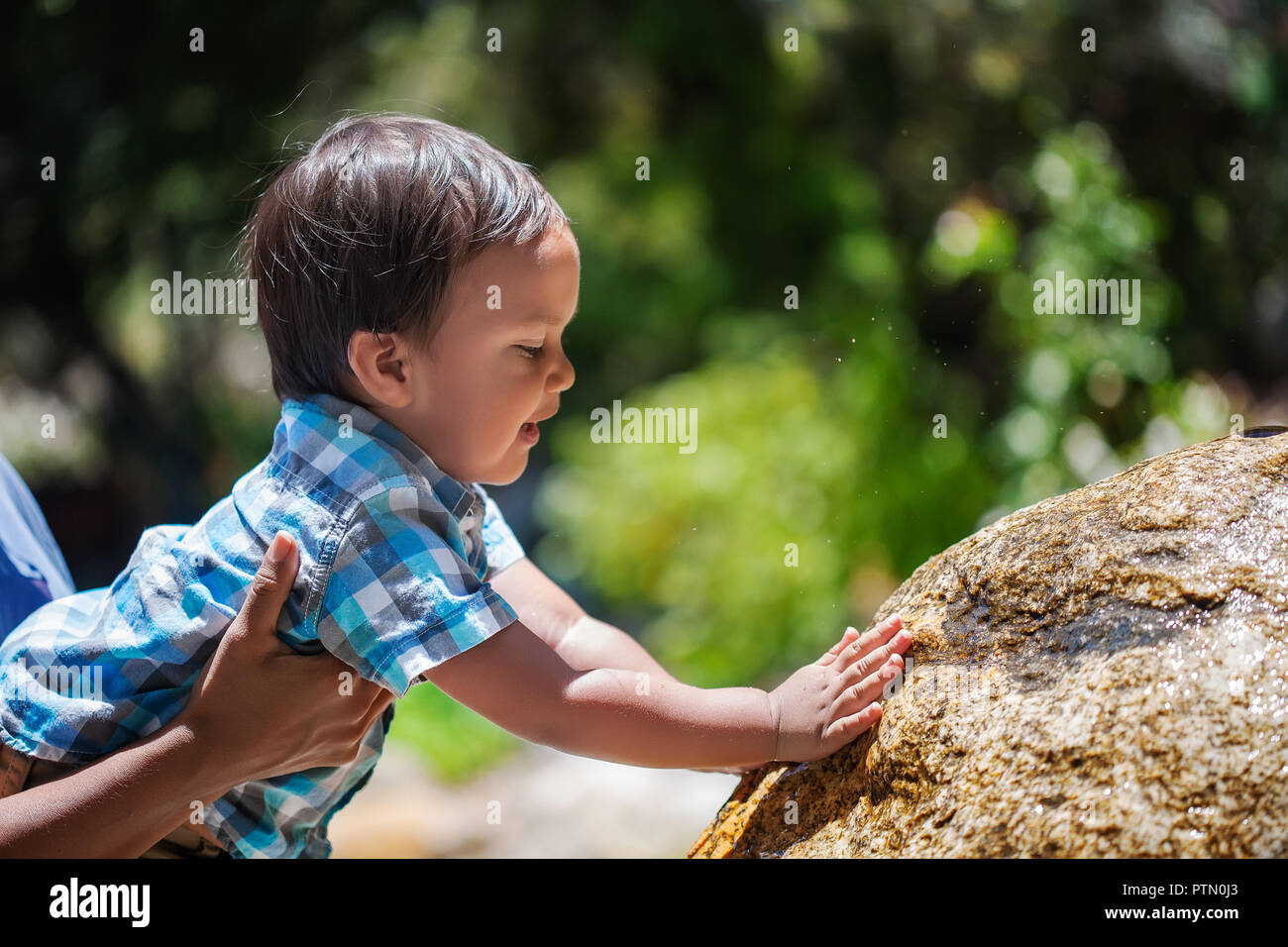 Mother supporting her toddler boy as he explores touching a natural water fountain outdoors and splashing Stock Photo