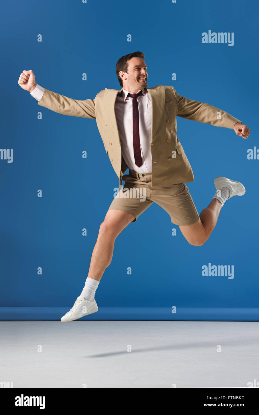 excited man in shorts and suit jacket jumping and looking away on blue Stock Photo