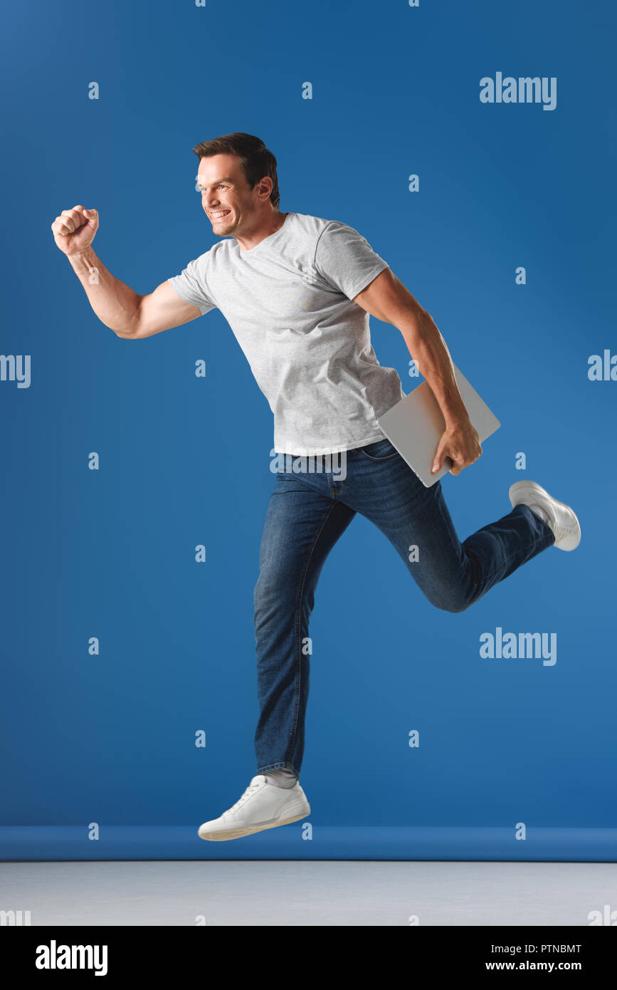 cheerful man holding laptop and jumping on blue Stock Photo