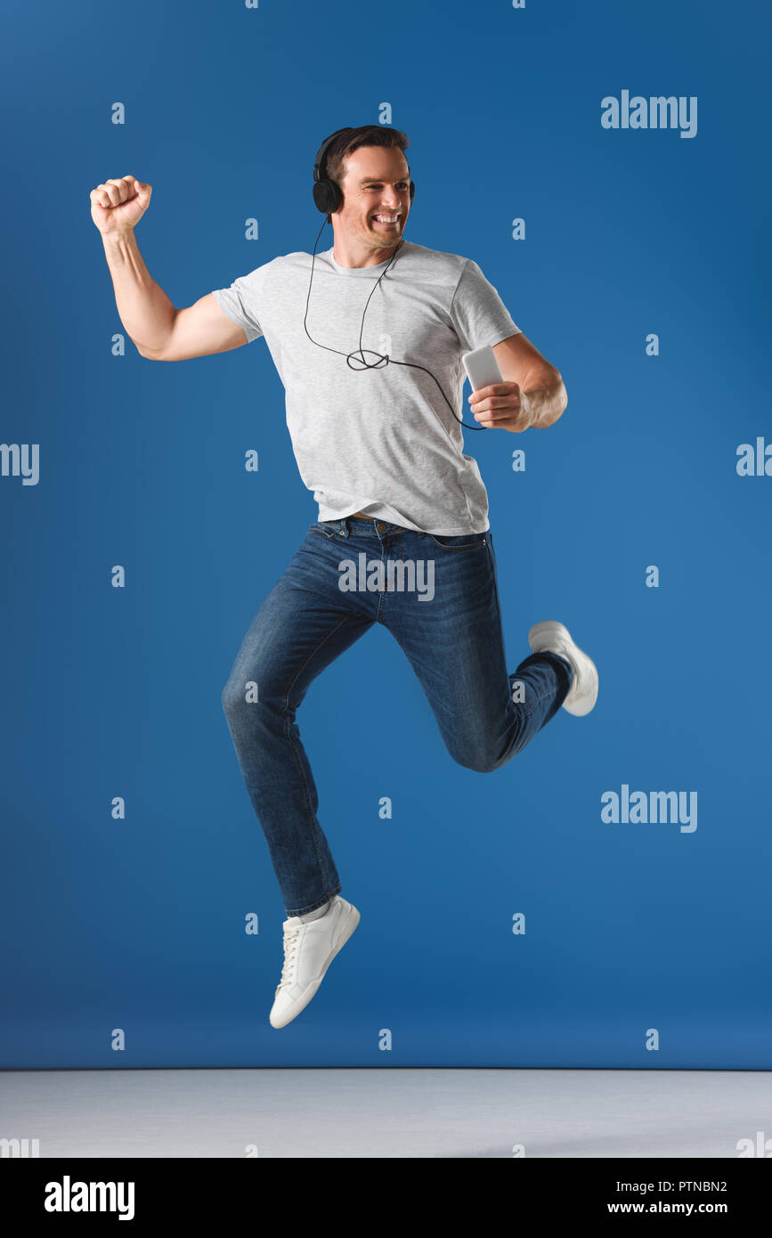 cheerful man in headphones holding smartphone and jumping on blue Stock Photo